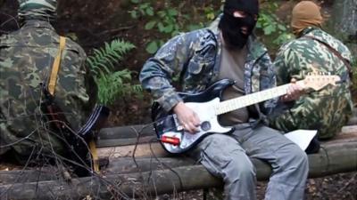 Masked men in military uniforms, one playing a guitar.