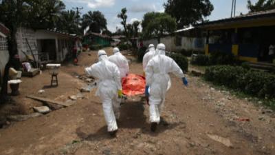 Four health workers in protection suits carry a body in an orange body bag