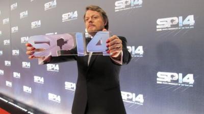 Eddie Izzard standing in front of a wall with SP14 on it, holding a glass SP14