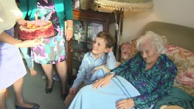 Gladys Hooper receiving her birthday cake, with a great-grandchild by her side