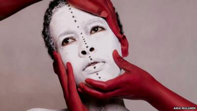 Photograph by Aida Muluneh of an African woman, her face painted white face and being held by three hands, painted red