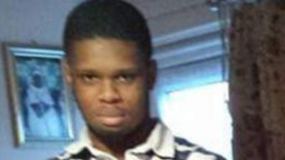 A blurry home photo showing Ahmed Deen-Jah. He is standing in front of a window in what appears to be a living room and is looking at the camera. He is wearing a top with a collar