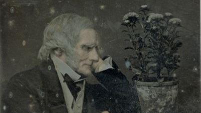 The image shows an elderly man with grey hair wearing a suit and looking at a pot of flowers.