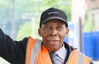 Siggy Cragwell, who was awarded a British Empire Medal for his long service on the railways, waves on a station platform. He is wearing a Thameslink railway cap and a fluorescent orange jacket, a shirt and tie.