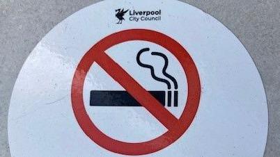 A "no smoking on the street" sign, featuring Liverpool City Council's emblem, has been stuck to the ground.