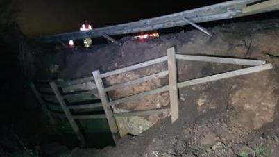 Bent crash barriers along the top of a bank of earth, with a broken wooden fence on the side of the bank. Traffic headlights appear in the background.