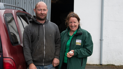 Shows a burgundy-coloured vehicle to the left then Vincent in blue jeans and a grey zip-up hooded to and Annie in a green T-shirt and darker green top, holding two eggs in her left hand