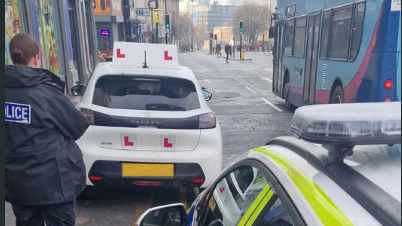 Police officer taking notes on the parking of the white learner car covered with L plates, which is parked partly on the pavement over double yellow lines