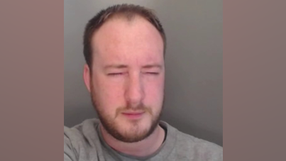 A custody photo of Nathan Henderson who has short brown hair and a beard and is wearing a grey shirt. His eyes are closed and he is leaning to the left of the frame.