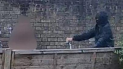 A view over the fence into a street or alleyway. A man on a bike is riding, there is a wall on the far side. The rider is wearing all black with a black balaclava.