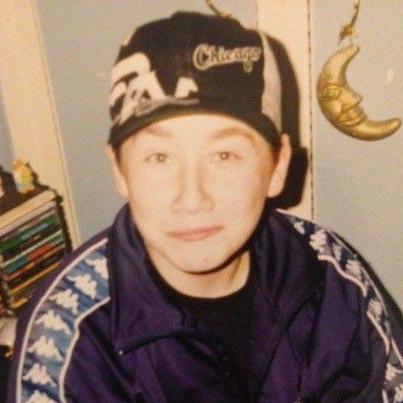 A blurry old photo of Stuart Ralston as a young teenager - in a navy Kappa tracksuit top and a backwards "Chicago" cap. A stack of CDs sit behind him and a gold moon ornament is hanging on the wall on the other side.