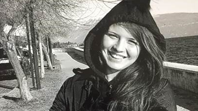 A black and white photo of the late Natasha Melendez.  She has long, straight hair and is smiling at the camera.  She is wearing a dark, hooded coat.  She is standing on a tree-lined path with a wall and a large body of water behind her. 