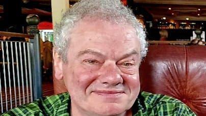Brad smiling and wearing a green plaid shirt sitting in a leather chair. A bar can be seen behind him. He has short silver curly hair