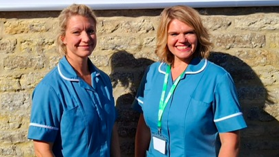 Zoe Draper and Gemma Crane are wearing blue nurse's uniforms and have blonde hair. Both are smiling. They are stood in front of a sign that says "The Hummingbird Centre: cancer support and therapy centre"