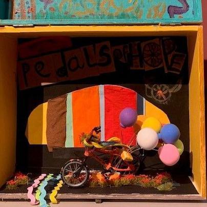 A wooden box, with its lid flipped open, depicts Nahla's scene of hope - a tiny model of an orange bicycle with coloured  balloons streaming behind it. The background is cheerfully-coloured stripes of fabric. The box is pained yellow and the lid is painted in happy colours.