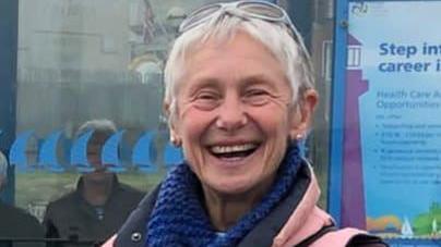 Dilys Gartside smiles broadly in a street outside an office. She has sunglasses pushed back over her short, white hair and wears a pink gilet and blue scarf.