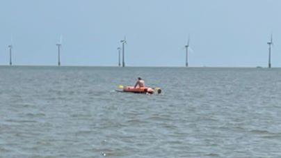 An inflatable boat is in the middle of the sea with a person on it. In the background there are six wind turbines in the water.