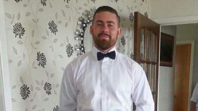 Frazer Irvine stood in a room which has flowery wallpaper. He has a brown beard and short hair. He is wearing a white shirt and black bowtie. A wooden door behind him is open.