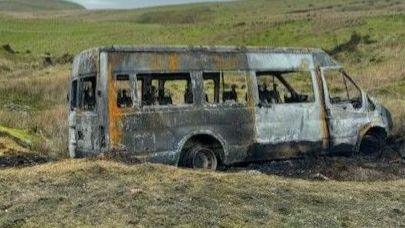 A torched school bus on a bank with green hills seen behind it. A tyre can be seen behind the vehicle, which is blackened with no wheels. A curving road can just be seen out of shot on the left hand side.