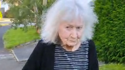 Anne Stacey walking down a pavement. The picture is from the waist up. She has grey bobbed hair and is wearing a striped navy blue and white top and a black cardigan. Driveways, lawns and trees can be seen in the background