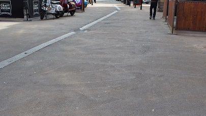 Close up of road with cracked concrete and darker grey spots. There is a person walking in the distance.
