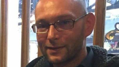 A man wearing glasses is smiling at the camera. His head is shaven, and he has stubble.