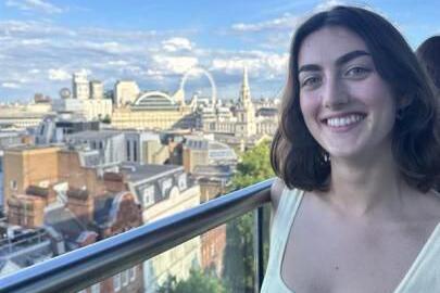 Eleanor Wallis is wearing a white top and is smiling at the camera. There is a view of London on a partly cloudy day behind her.
