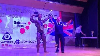 Mr Rosiek poses on a stage with arms raised, wearing only trunks and a medal around his neck whilst another man is standing next to him wearing a waistcoat and trousers with a shirt and tie whilst holding a Union Jack flag over his shoulders.