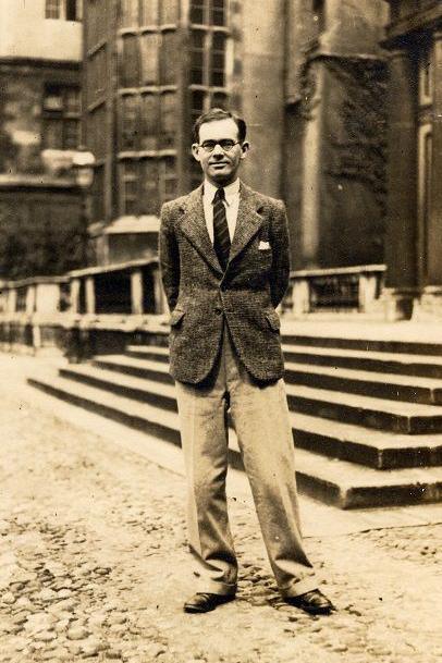 Student Gareth Jones at Cambridge University, with his hands behind his back, standing in front of the steps leading to a university building. It is a black and white image, but more a sepia tone. He has a blazer on, and a white shirt and tie and trousers. He wears rounded glasses.