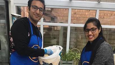 Volunteers in the kitchen. A man wearing a blue apron and hygiene gloves hold a bowl in his hands next to a woman who also wears a blue apron. Both are smiling at the camera. 