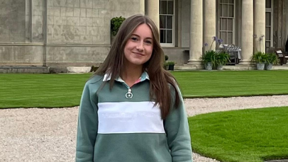 Bethany is standing near an old stone building. She is smiling and wearing a green and white jumper. Her long brown hair is down.