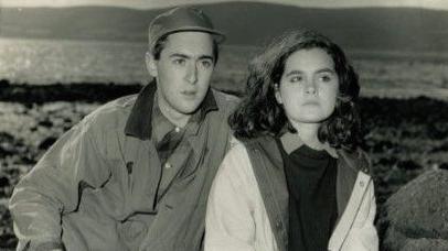 A black-and-white photograph shows Allan and Shirley seated among large rocks on a shoreline, with water and distant hills visible in the background.
Both are dressed in outdoor clothing, creating a candid, natural scene in a rugged coastal setting.