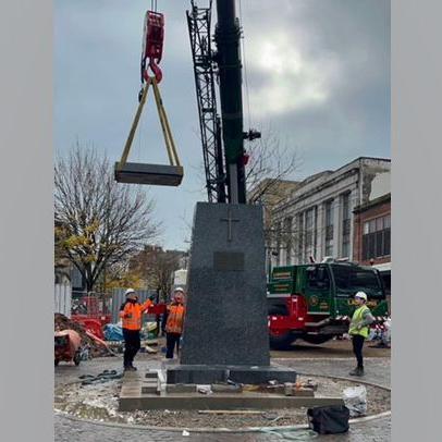 Workman moving the memorial. Three men in hi-vis jackets are guiding a small crane. The crane is lowering the top pyramid of the obelisk into position.