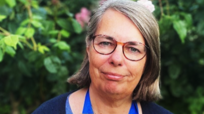 A woman, with short grey hair, wearing glasses. She is smiling at the camera. She is wearing a blue top and cardigan. There is a green bush behind her. 