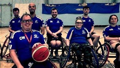 A wheelchair basketball team wearing blue sports tops and black shorts are smiling at the camera. Four men are on the back row and two women and one man are on the front row. 