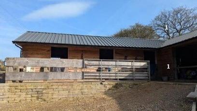 The image shows a picture of the existing barns. The barn is made of brow wood and have a black slanted roof. There is a brown wooden fence covering part of the entrance to the barn. The barn is built onto a brick platform.