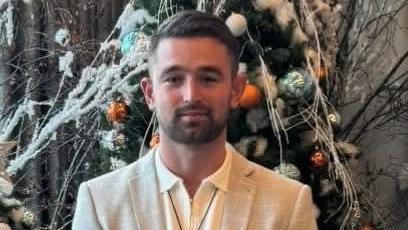James Reynolds, who has dark hair and a short dark beard, is wearing a cream suit jacket over an open-collar cream shirt and is standing in front of a tree bedecked in white, blue and orange baubles. 