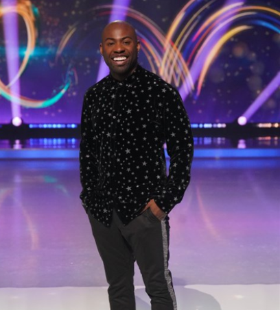 A man in a black sparkly shirt and grey striped trousers smiles at the camera.