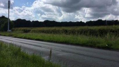 A stretch of road surrounded by a green, grassy fields with a line of trees visible in the backdrop and a cloudy sky above. 