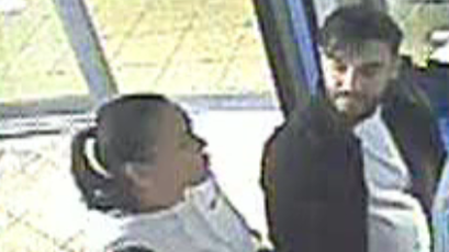 CCTV from the entrance of a bus, pictured on the left is a woman with dark hair pulled back into a ponytail, wearing a white jacket. On the right is a man with dark hair and short fringe, he also had a beard and moustache. He is wearing a white t-shirt and dark jacket.