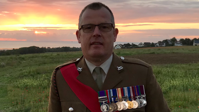 Paul Smales, wearing his ceremonial dark green Army uniform, is pictured against a sunset sky. He has nine medals on his chest and a red sash across his body. 