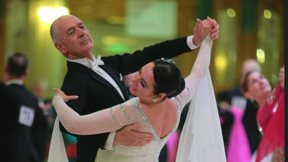 A man and woman in formal dress are dancing together. The man wears a black suit and cream bow tie, the woman is in a cream sparkly dress, with flowing material