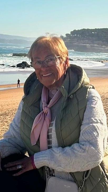 Maureen Harris smiles at the camera as she stands near a beach. She has short blonde hair and is wearing a thick knitted jumper underneath a green gilet.