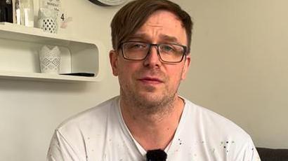 Gregory Lesiak sitting on a chair looking into a camera. He has dark hair and is wearing glasses and a white t-shirt.
