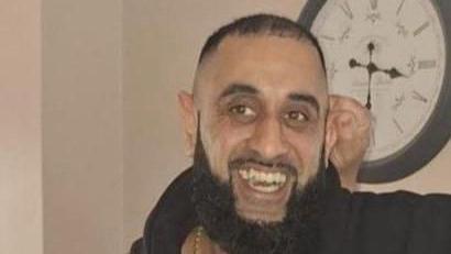 Mohammed Osman Khan wearing a black hoodie stood in front of a wall with a clock 