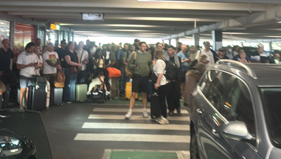 Dozens of people stand in a car park by a zebra crossing with their luggage in among parked cars.