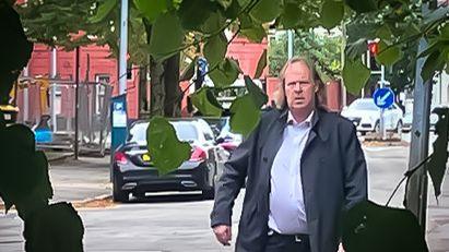 older man with long hair balding on top, wearing an open jacket with a large stomach sticking out