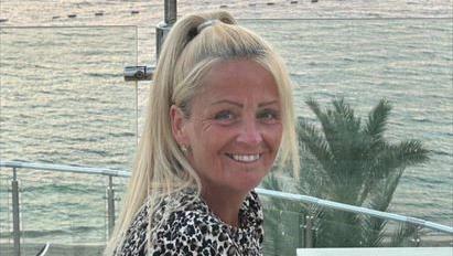 Anita Rose is wearing an animal print top and sat in front of a railing by the sea. Her blonde hair is in a high ponytail and she is looking directly at the camera and smiling. In the background is a palm tree and the sea.