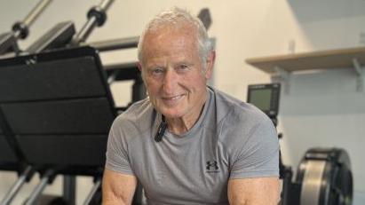 Peter is wearing the a grey gym t-shirt, and is smiling for the camera as he sits on a weight lifting bench. There are exercise machines behind him and to the left of him.