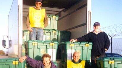 Four men sit and stand around green plastic boxes. They are position at the back of an open lorry. Two men are wearing bright yellow bibs. 